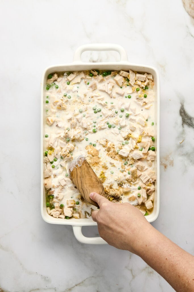 A hand stirring a creamy chicken and green pea orzo casserole in a white baking dish, garnished with chopped herbs, on a marble countertop.