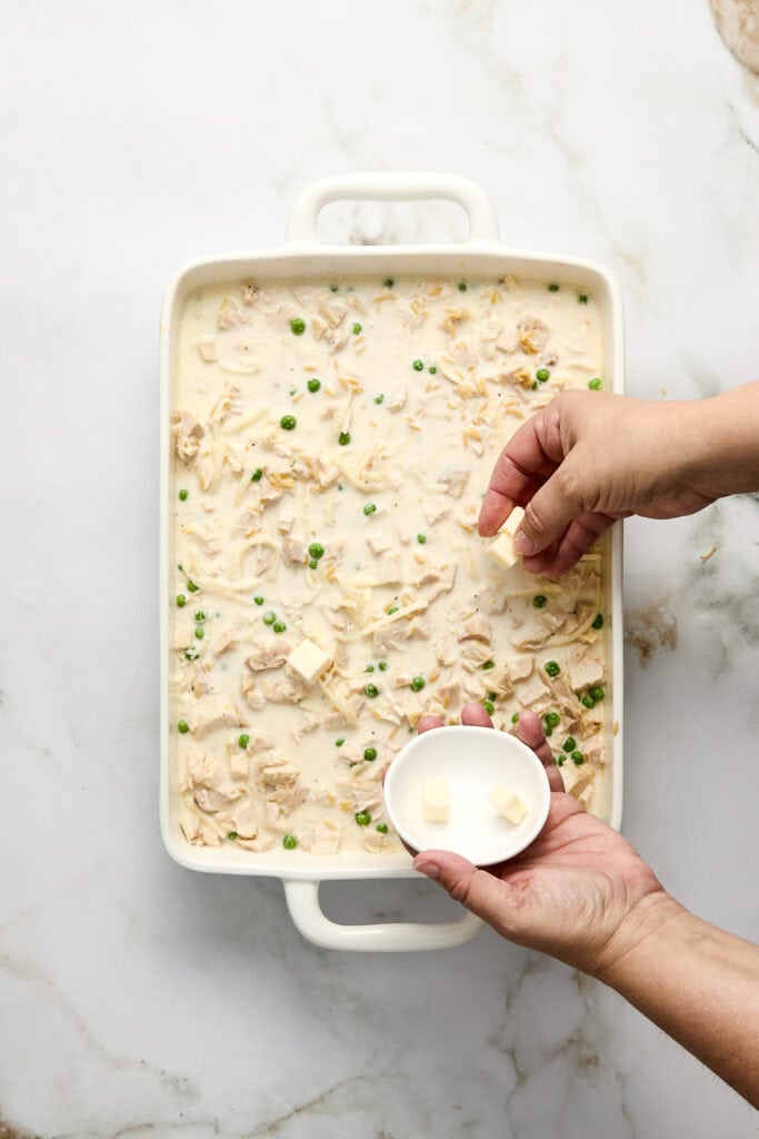 Close-up of a creamy chicken and pea orzo casserole in a white baking dish, with a hand adding cheese cubes on top, on a marble surface.