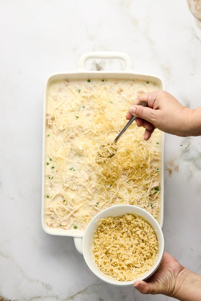 Close-up of a creamy casserole dish topped with shredded cheese and green herbs, with a hand sprinkling more cheese from a bowl, on a white marble surface.