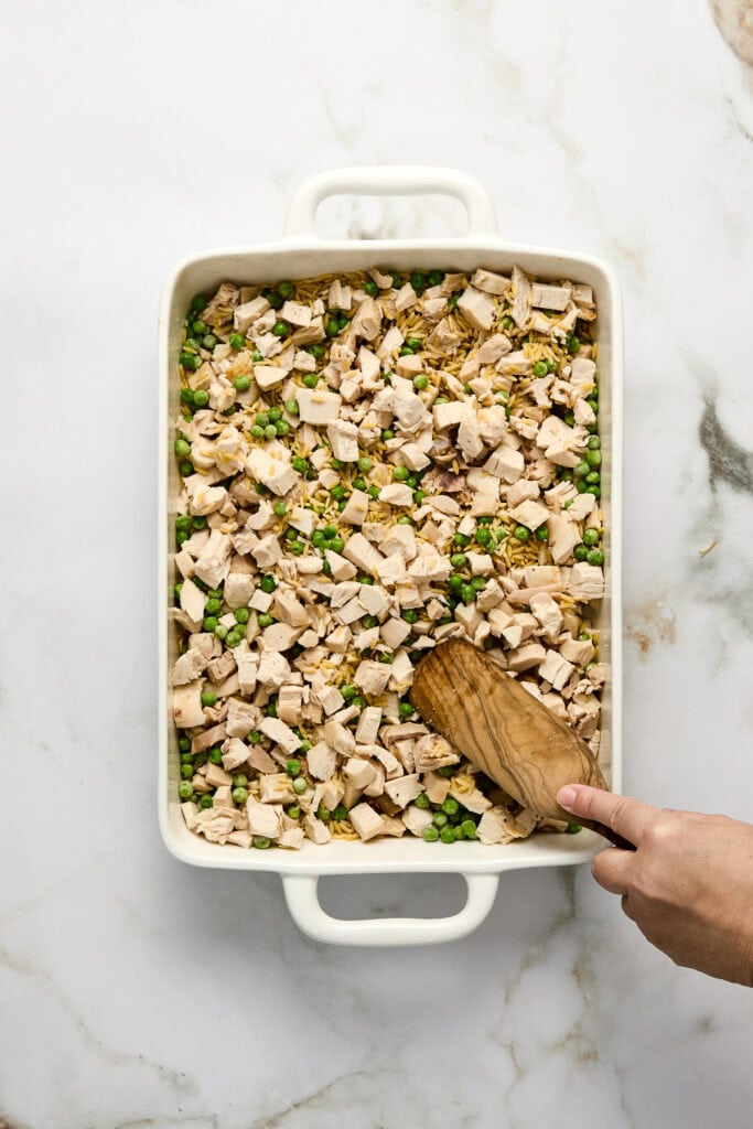 Baked chicken orzo and green peas casserole in a white baking dish, with a hand using a wooden spoon to stir the ingredients.