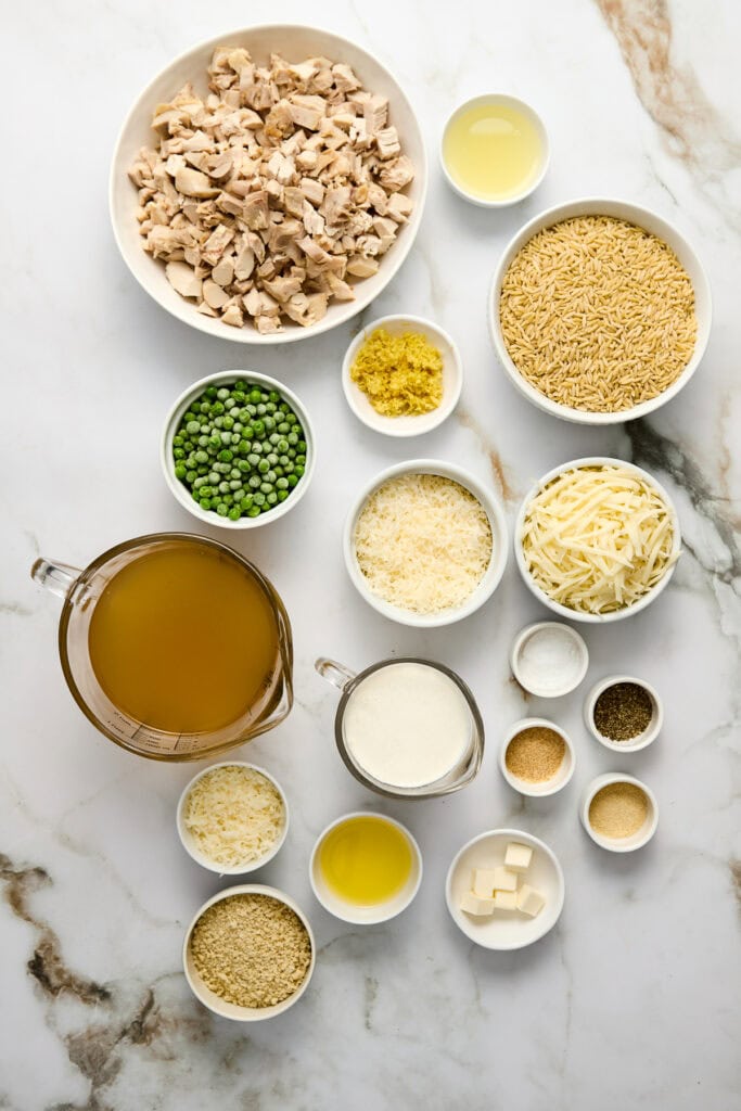 Assorted ingredients for creamy chicken and pasta dish including cooked chicken, pasta, peas, cheese, and seasonings arranged on a marble surface.