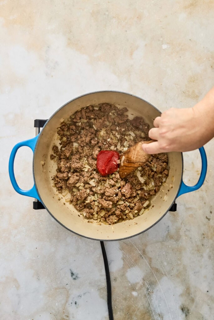 Person stirring ground beef and tomato sauce in a blue skillet on a light-colored surface.