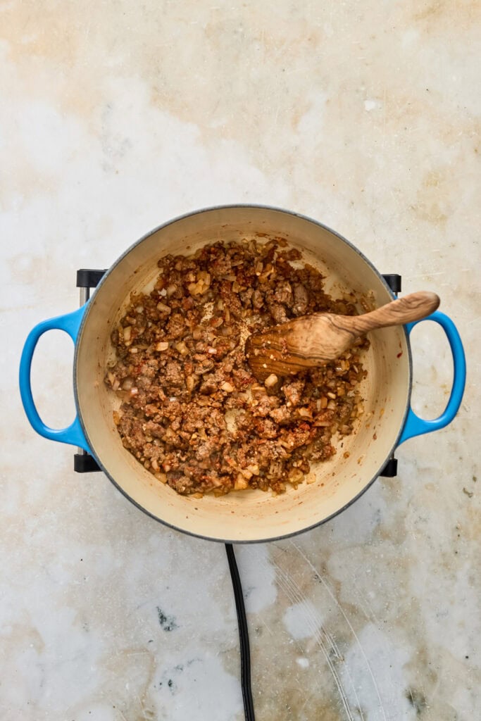 Ground meat being cooked in a pot with a wooden spoon, on a light-colored surface.