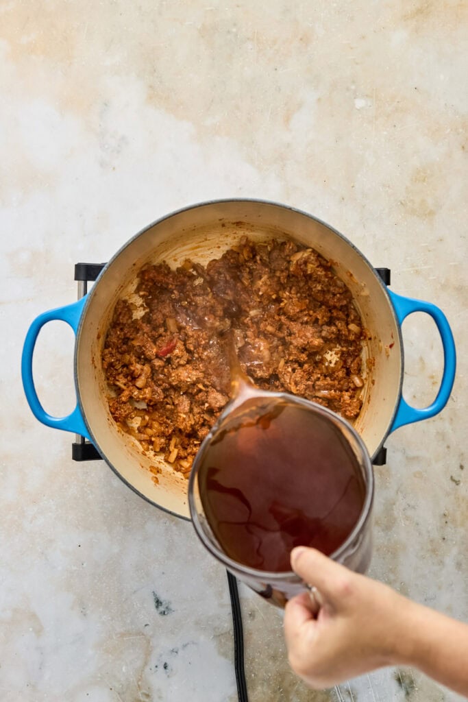 A pot of cooked ground beef with tomato sauce on a stove, with a hand pouring a glass of red wine into the pot.