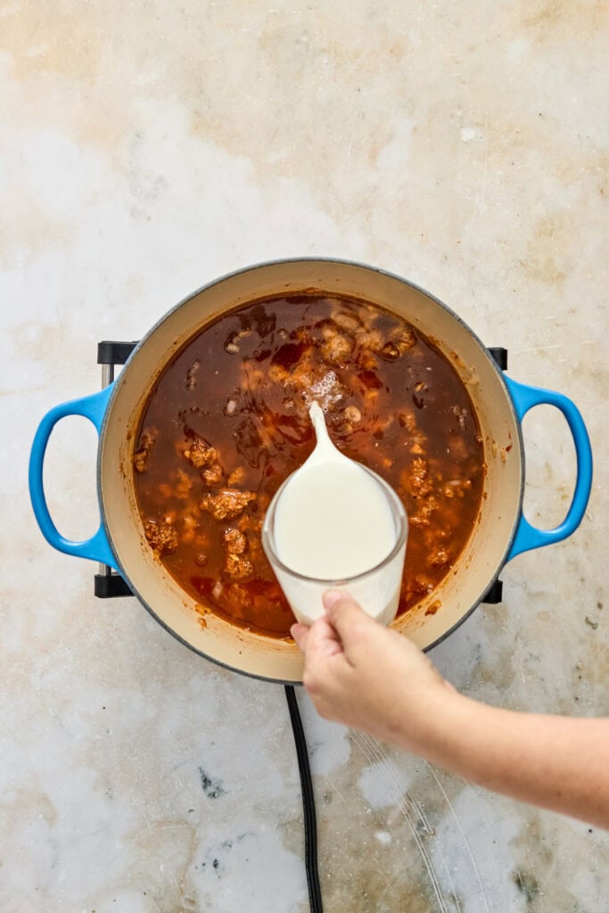 A person pouring cream into a spicy sauce in a large blue pot, top-down view, on a light textured surface.
