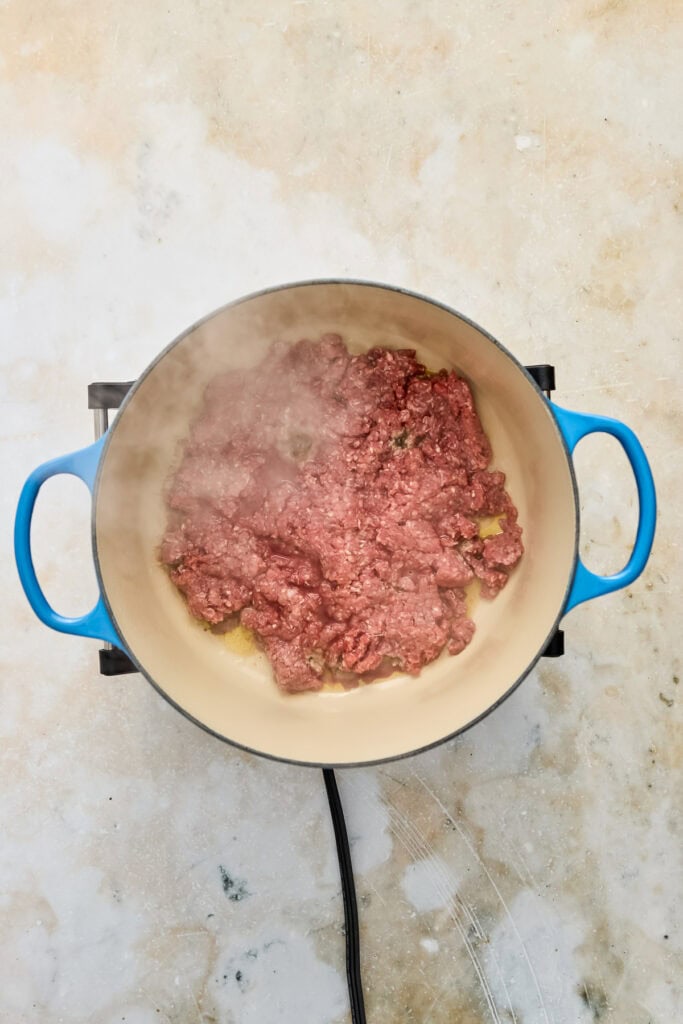 Ground beef being cooked in a blue pot on a stovetop, with steam rising from the meat.