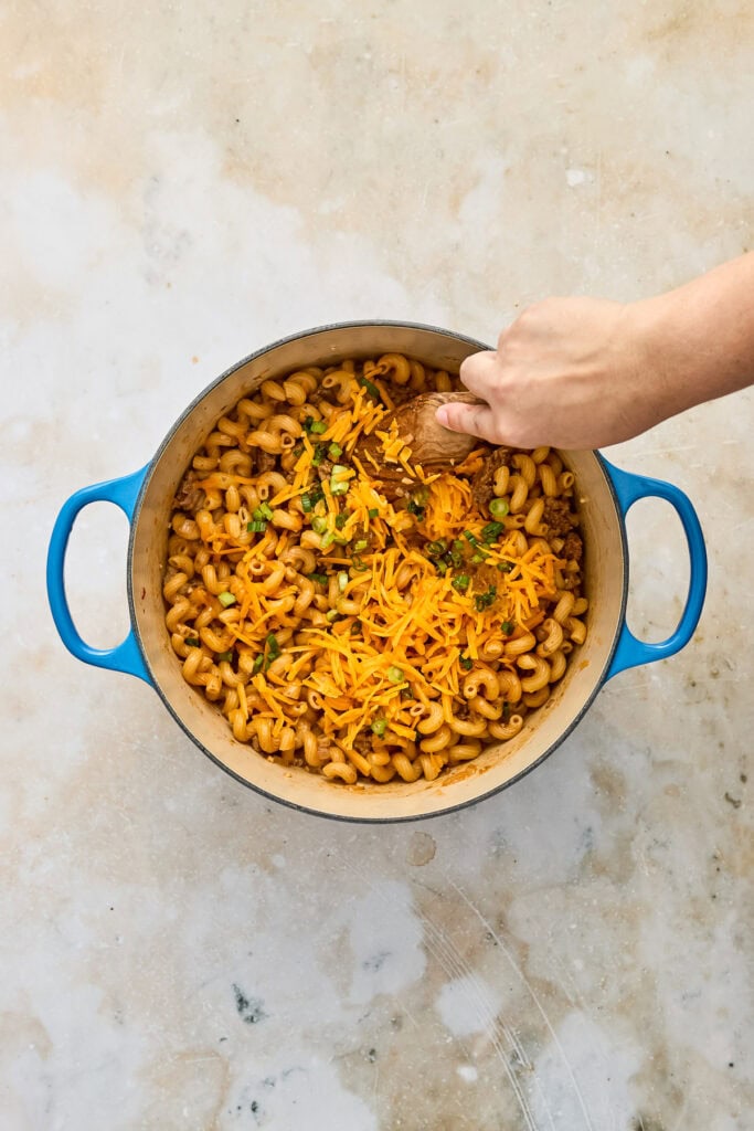 A hand stirring a cheesy pasta dish with shredded cheese and herbs in a blue pot on a neutral surface.
