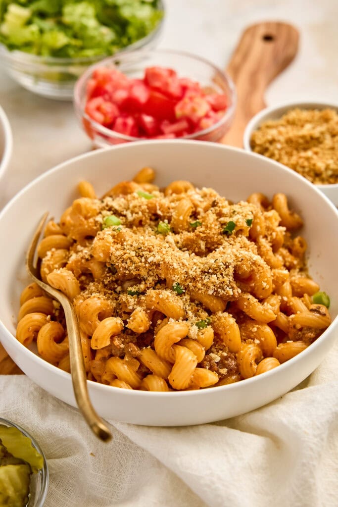 Close-up of a bowl of creamy cheeseburger pasta with breadcrumb topping, garnished with green herbs, served with fresh salad and diced tomatoes in the background.