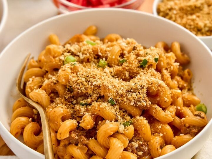 Close-up of a bowl of creamy cheeseburger pasta with breadcrumb topping, garnished with green herbs, served with fresh salad and diced tomatoes in the background.