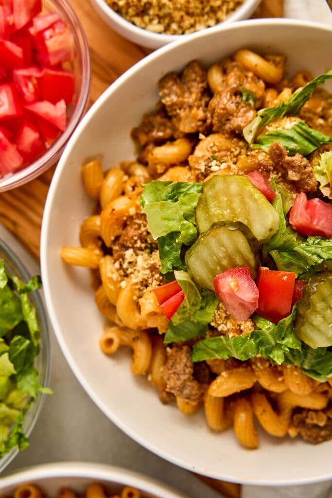 Close-up of cheeseburger pasta with ground beef, lettuce, pickles, and diced tomatoes in a white bowl, with side dishes of watermelon and greens.