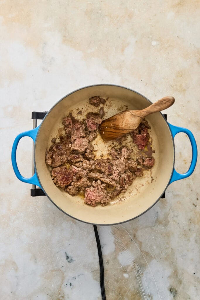 Ground beef being cooked in a blue pot with a wooden spoon, on a light-colored surface.
