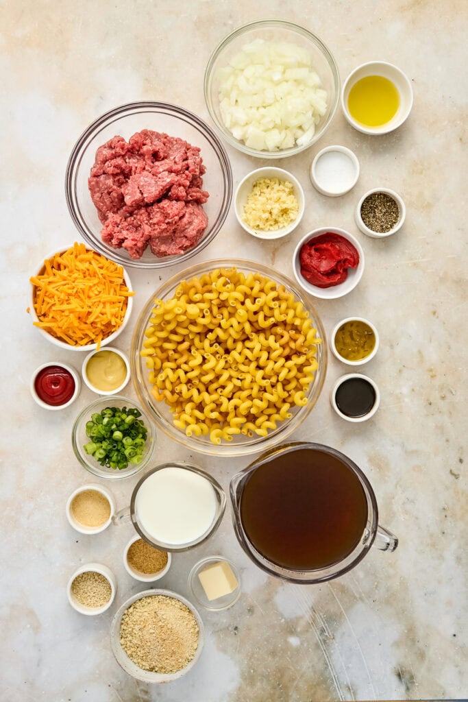 Assorted ingredients for making a flavorful pasta dish, including ground meat, pasta, cheese, and spices, arranged on a light surface.