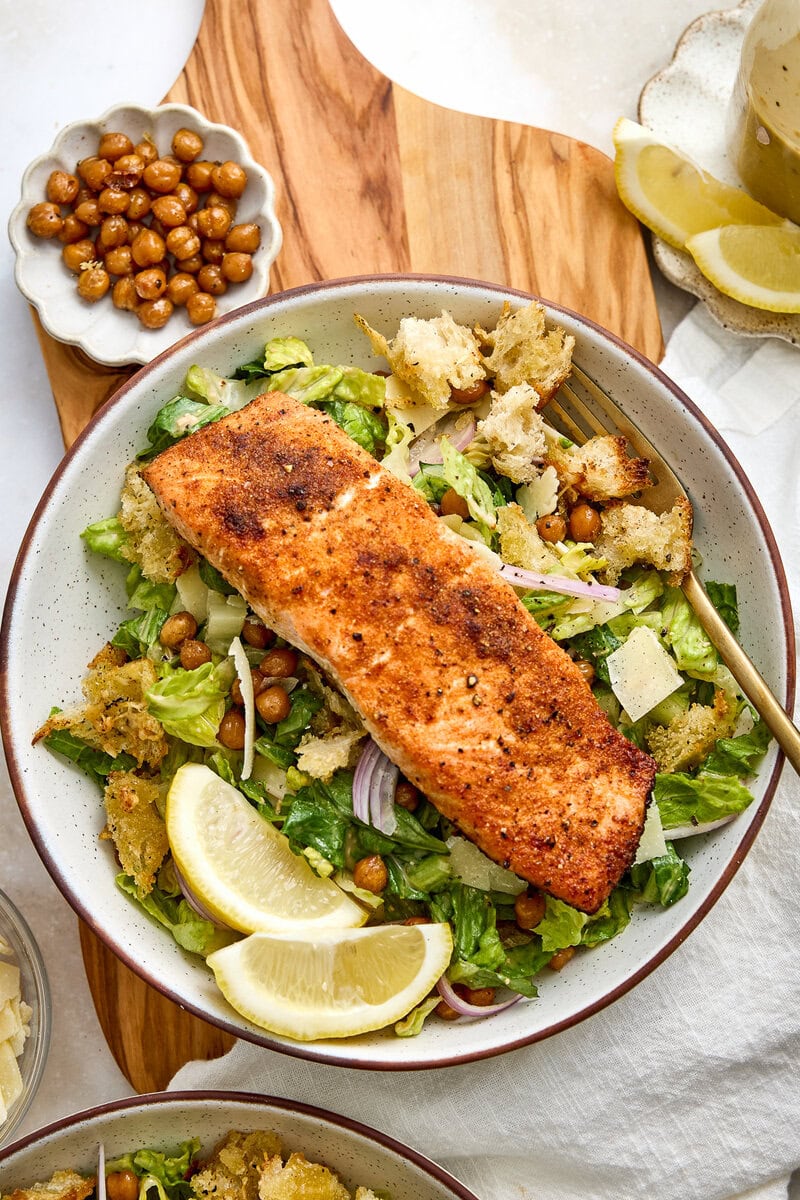 Delicious crispy salmon Caesar salad with roasted chickpeas, garlic Parmesan croutons, lemon wedges, and fresh greens, perfect for a healthy meal.