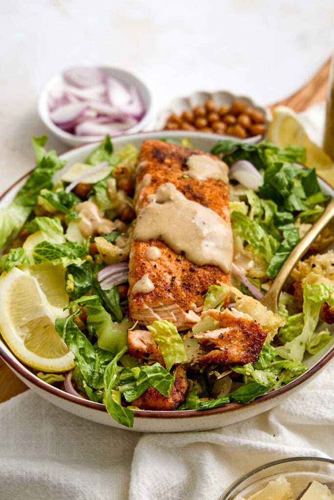 Fresh salmon caesar salad with mixed greens, lemon slices, and creamy dressing, served with side dishes of chickpeas and sliced onions.