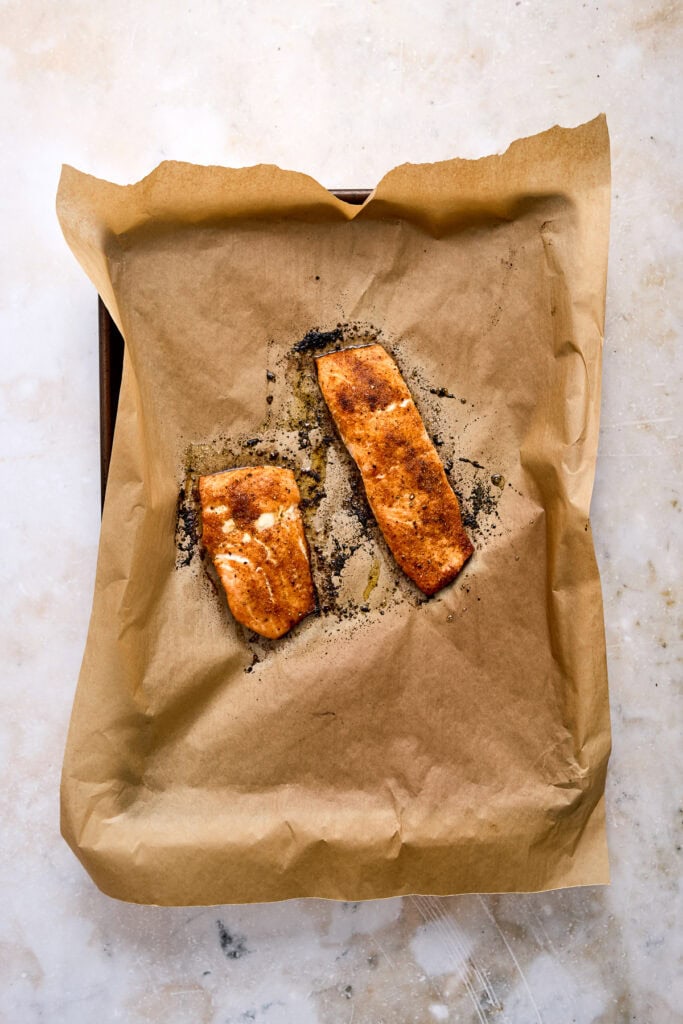 Two baked salmon fillets with crispy skin on brown paper, garnished with herbs, on a light marble surface, perfect for healthy meal ideas.