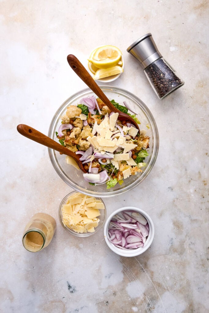 Fresh Caesar salad with crispy salmon, roasted chickpeas, garlic Parmesan croutons, and sliced red onions in a glass bowl, with lemon, pepper grinder, and dressing on a light background.