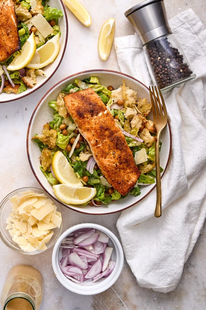 Delicious crispy salmon Caesar salad with roasted chickpeas, garlic Parmesan croutons, lemon wedges, and fresh greens on a white plate.