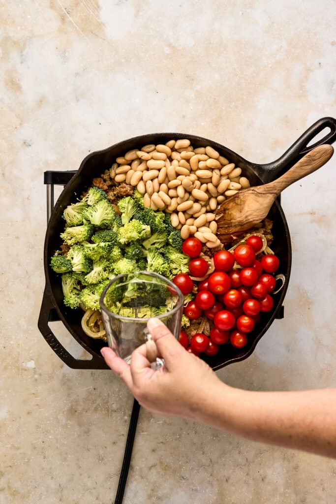Person adding water to a skillet filled with broccoli, cherry tomatoes, white beans, and pasta for a healthy vegetable sausage skillet.