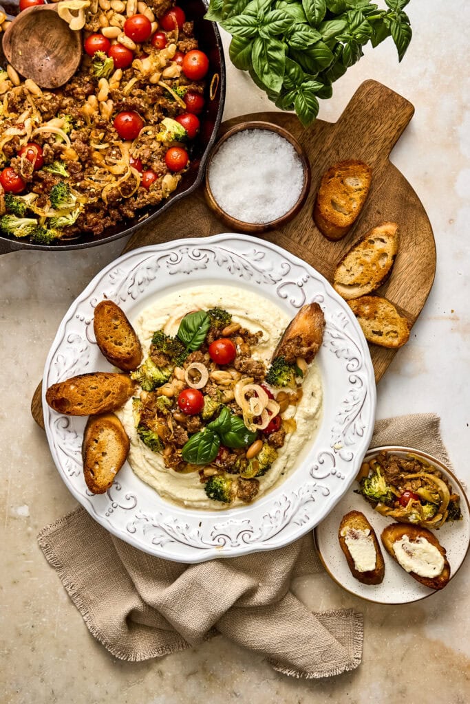 Colorful sausage and broccoli skillet served with toasted crostini, fresh basil, cherry tomatoes, and a side of salt in a rustic setting.