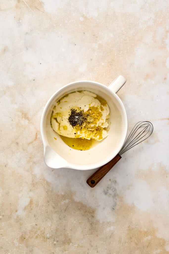 Creamy ricotta cheese with black pepper and lemon zest, with a small whisk nearby on a textured surface.