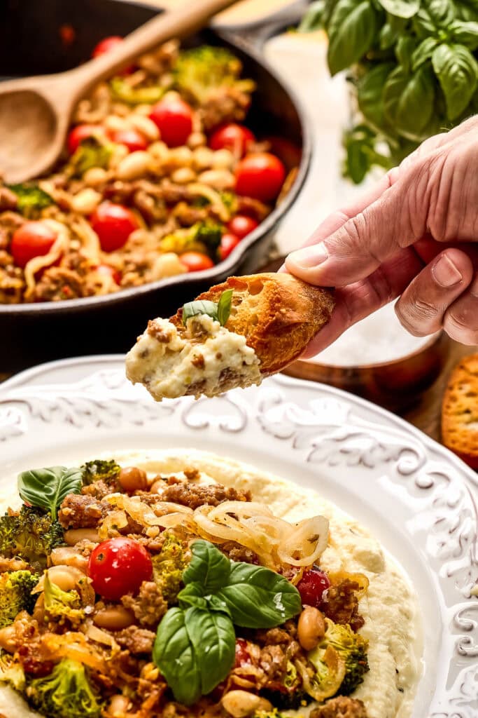 Close-up of a hand dipping a piece of bread into a flavorful lemon ricotta sausage and broccoli skillet with cherry tomatoes, chickpeas, and herbs on a decorative white plate.