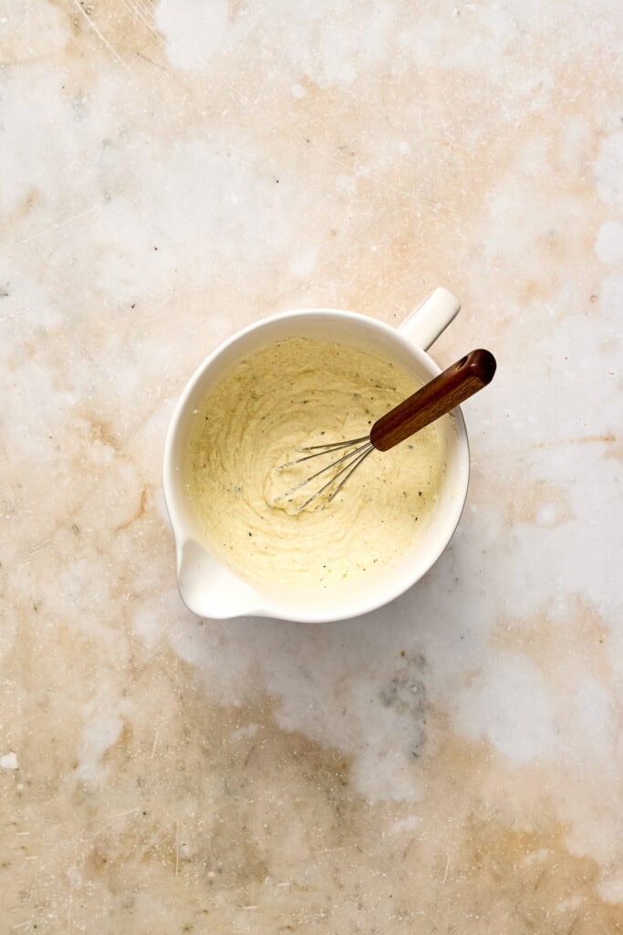 A white mixing bowl with creamy ricotta cheese and a whisk with a wooden handle, viewed from above on a textured surface.