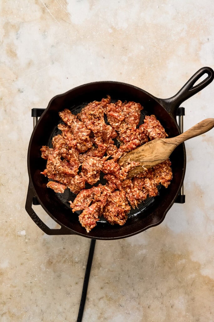 Ground beef being cooked in a black skillet with a wooden spatula on a light-colored surface.