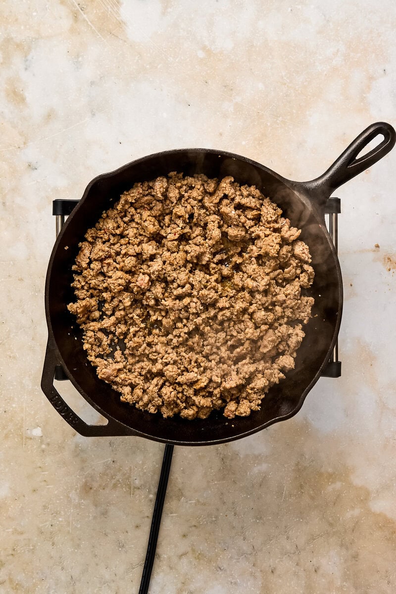 Italian sausage cooking in a black skillet on a stovetop, with a textured beige background.