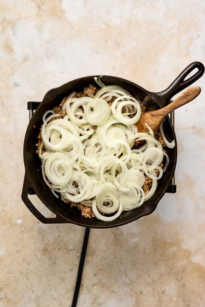 Sliced onions cooking in a cast iron skillet with a wooden spatula, ready for a savory dish.