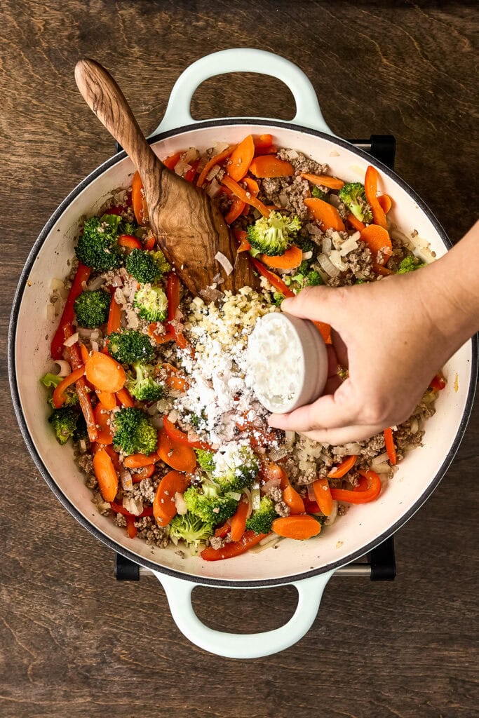 A hand adding grated cheese to a colorful vegetable stir-fry with broccoli, carrots, and ground beef in a white skillet on a wooden surface.