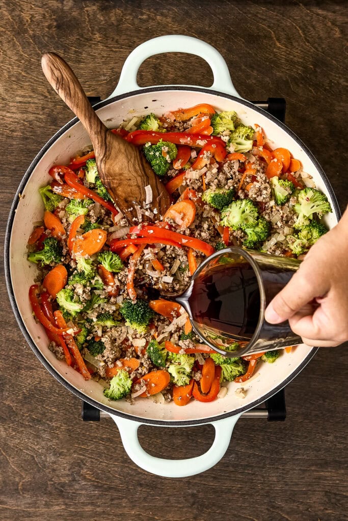 Colorful stir-fried vegetables including broccoli, red bell peppers, and carrots being cooked with soy sauce in a white skillet.
