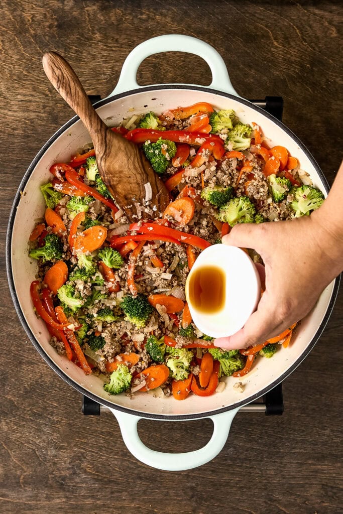 Colorful stir-fried vegetables including broccoli, red bell peppers, and carrots in a white skillet, with a hand pouring soy sauce.