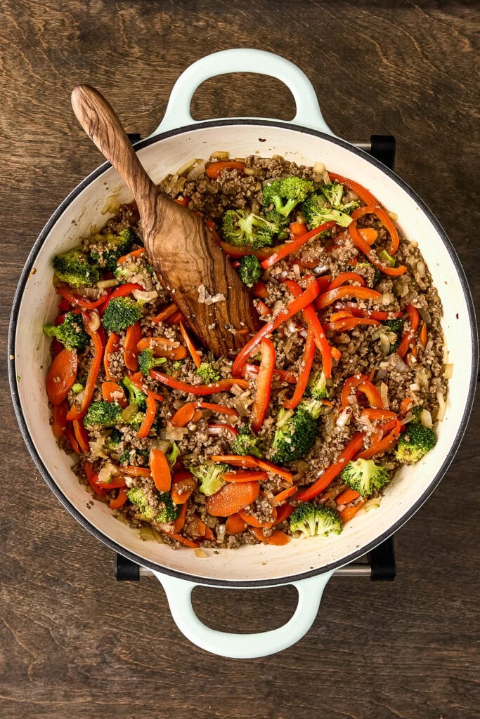 Colorful vegetable and ground beef stir-fry in a white skillet with a wooden spoon, on a wooden surface.
