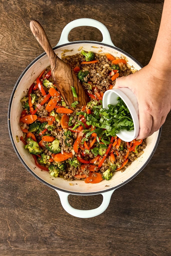 Fresh vegetable stir-fry with broccoli, carrots, and ground meat being prepared in a white cooking pot on a wooden surface.