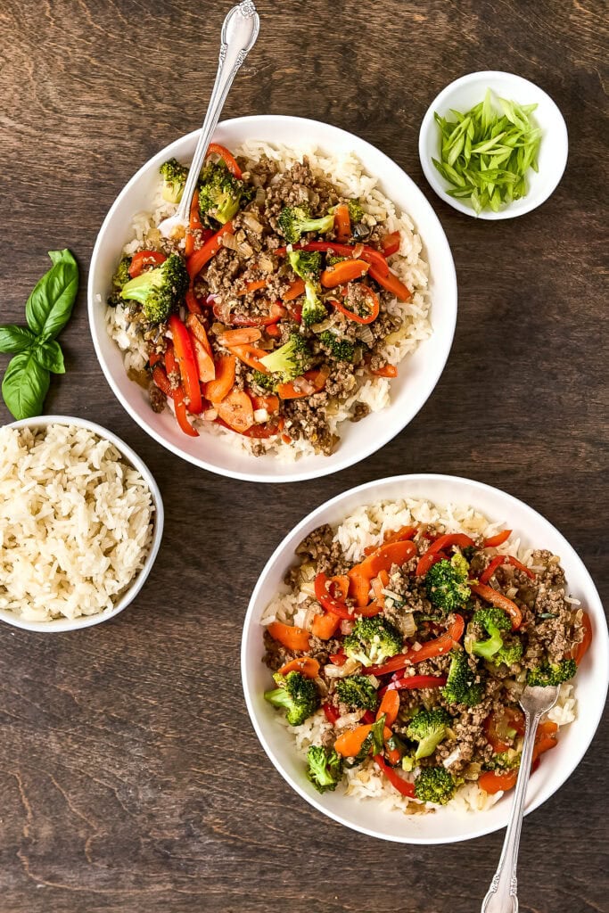 Colorful beef and vegetable stir-fry served in white bowls with rice, garnished with fresh herbs, on a wooden table.