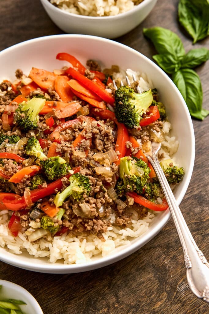 Colorful beef and vegetable stir-fry served over white rice in a white bowl, with fresh basil leaves on the side, perfect for a healthy meal.
