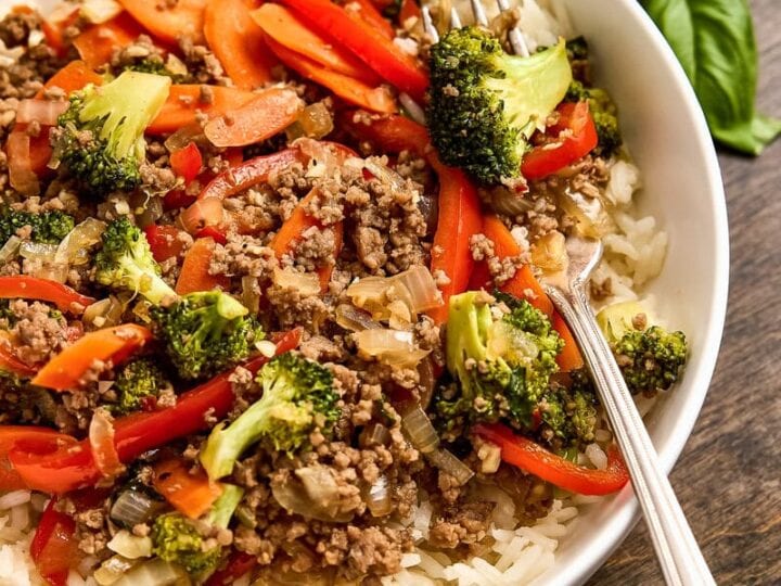 Colorful beef and vegetable stir-fry served over white rice in a white bowl, with fresh basil leaves on the side, perfect for a healthy meal.