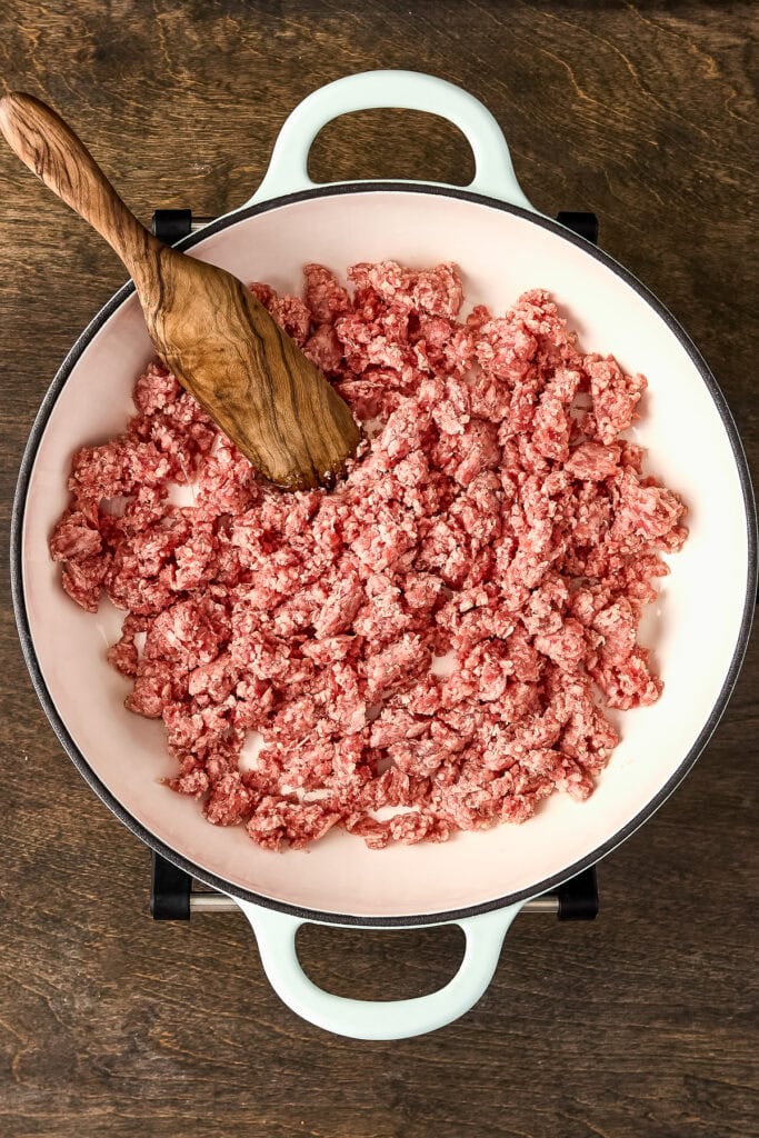 Ground meat being cooked in a white skillet with a wooden spatula, on a wooden surface, ideal for recipes and meal prep.