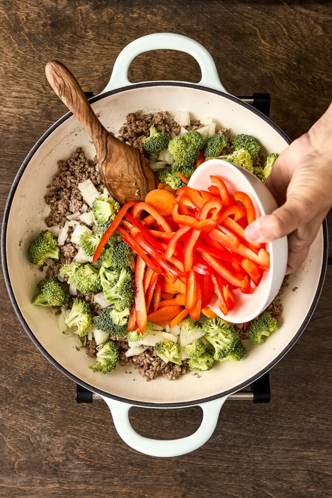 Fresh broccoli and red bell peppers being added to a stir-fry with ground meat in a white skillet on a wooden surface.