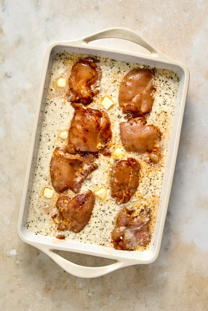 Baked chicken thighs in a white baking dish with seasoning and butter, ready for cooking or serving.
