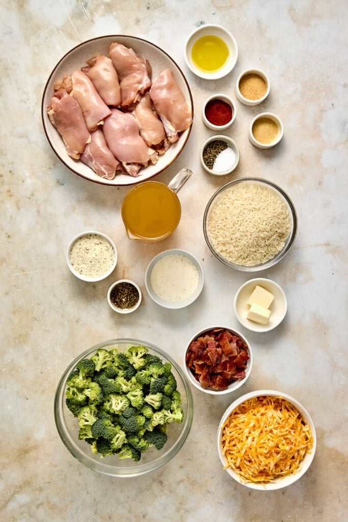 Raw chicken thighs, broccoli, rice, cheese, and spices arranged on a light surface for casserole preparation.