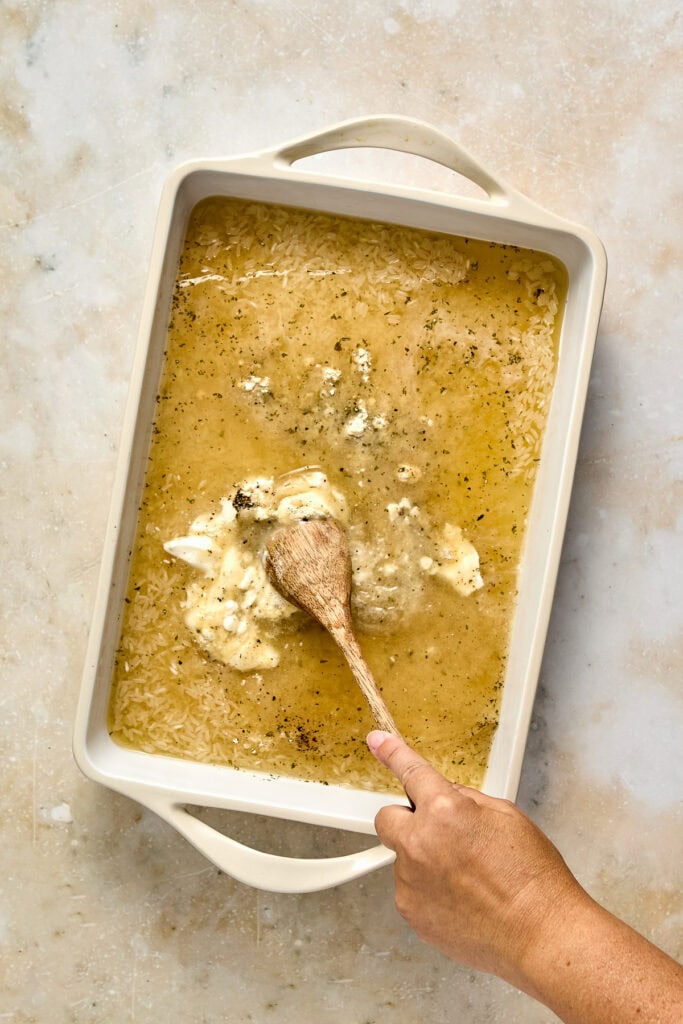 A person stirring homemade chicken soup in a white baking dish with a wooden spoon, on a light textured surface.