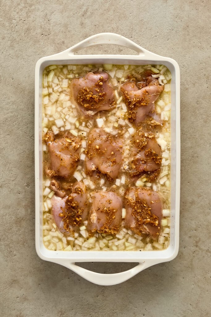 Raw chicken thighs in a baking dish with chopped onions and seasoning, ready for oven baking.
