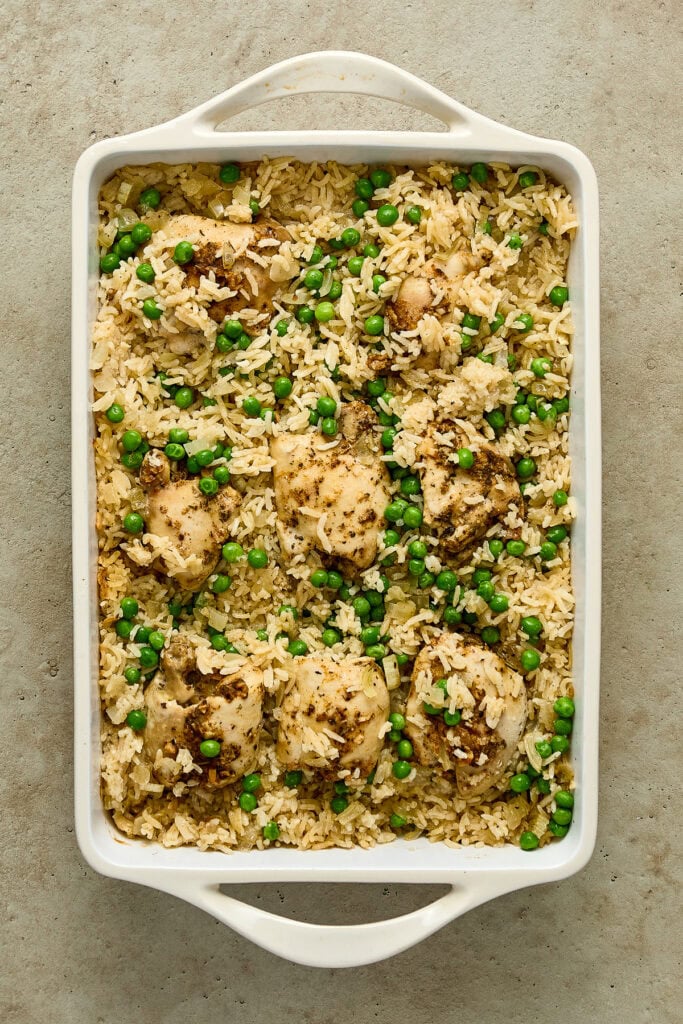 Baked chicken and rice casserole topped with green peas in a white baking dish on a gray surface.