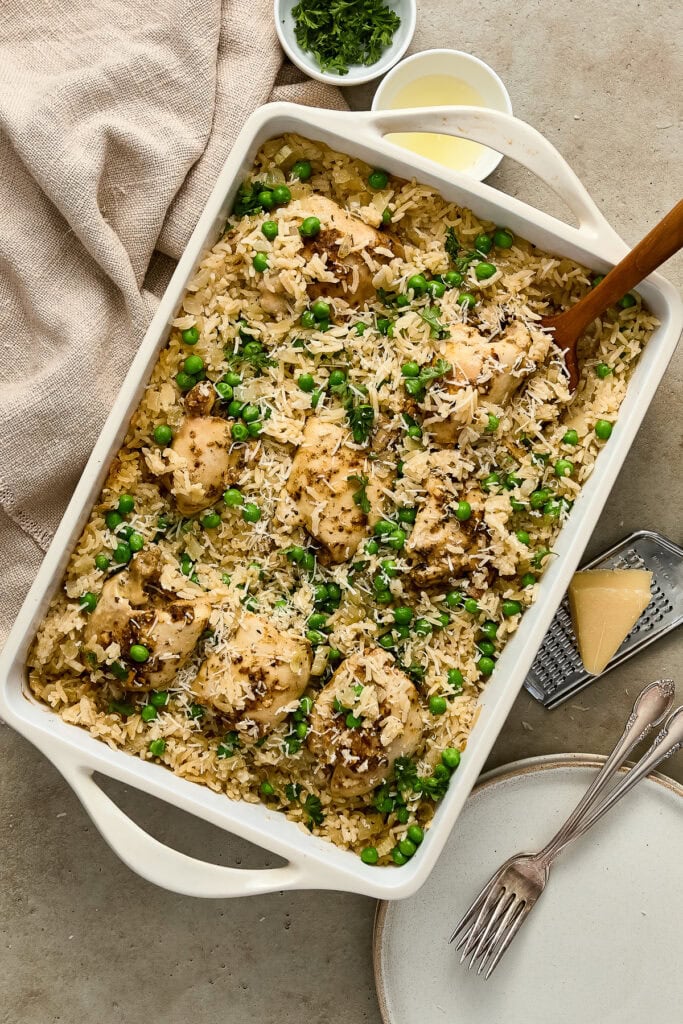 Delicious garlic chicken and rice casserole topped with green peas and herbs, served in a white baking dish with serving utensils.