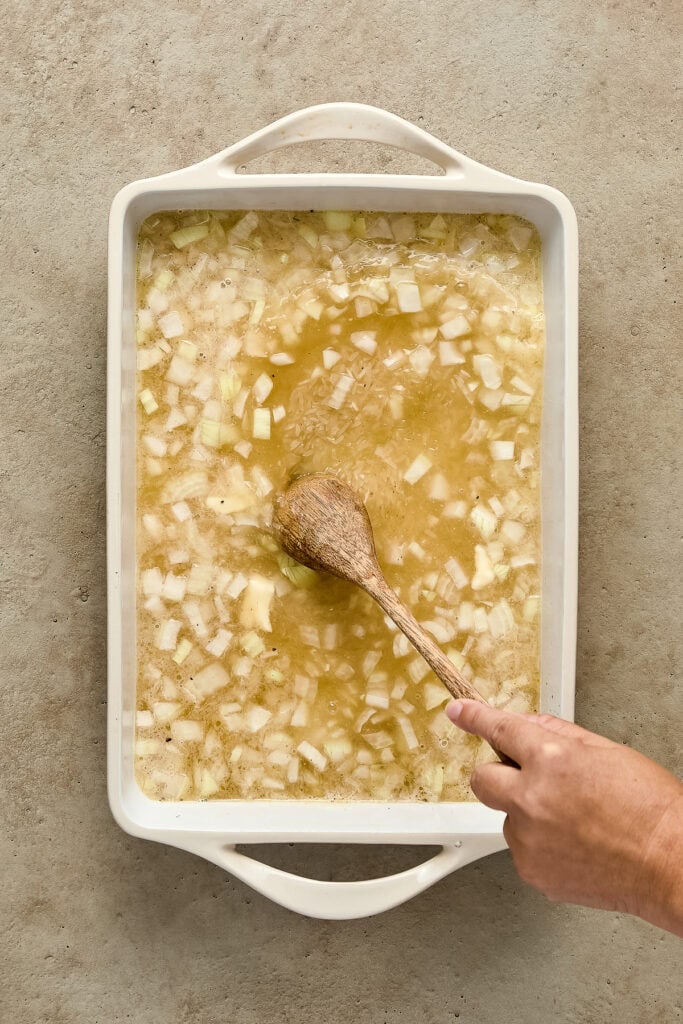 A person stirring a large tray of homemade onion soup with a wooden spoon, showing diced onions in broth.