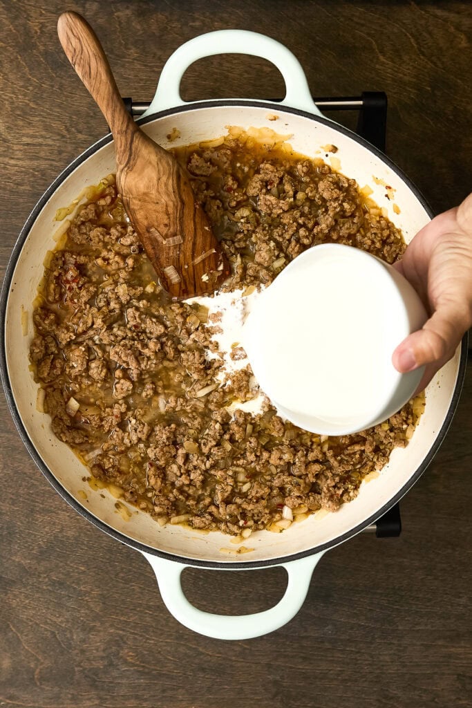 Cooking Zuppa Toscana Gnocchi with ground sausage, onions, and cream in a white skillet, wooden spoon stirring, on a wooden surface.
