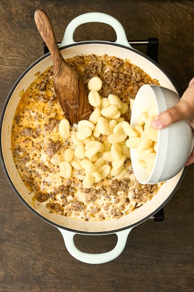 Creamy Zuppa Toscana with gnocchi, sausage, and kale in a white skillet, being prepared with cheese added from a bowl.