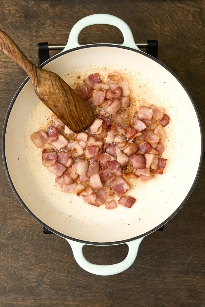 Bacon being cooked in a white skillet for Zuppa Toscana Gnocchi recipe, with a wooden spoon stirring the bacon.