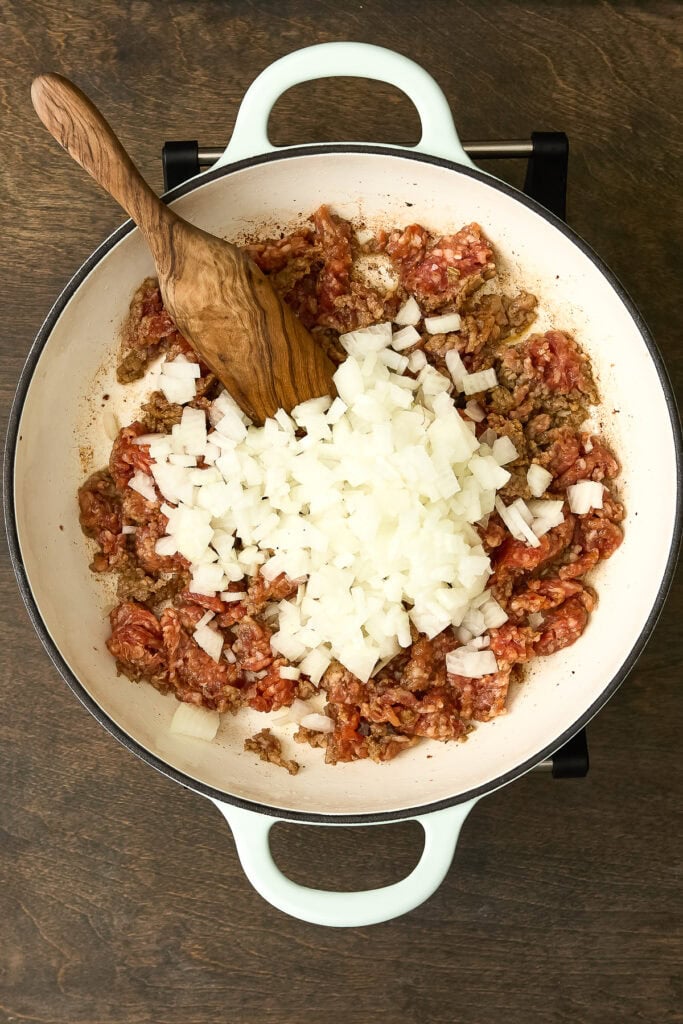 Preparing Zuppa Toscana Gnocchi with ground sausage, chopped onions, and tomatoes in a white skillet on a wooden surface.